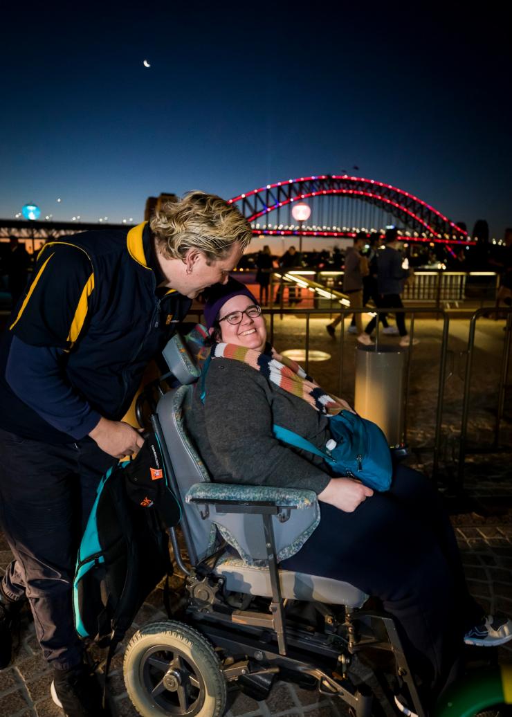 新南威尔士州，悉尼，缤纷悉尼灯光音乐节，一位女士坐在轮椅中，背后站着一个人，背景是悉尼海港大桥 © 新南威尔士州旅游局版权所有