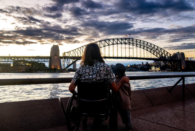 新南威尔士州，悉尼，环形码头，一位男子坐在轮椅上与儿子一同眺望悉尼海港大桥 © 澳大利亚旅游局版权所有