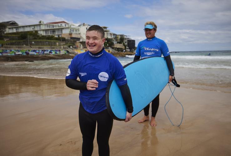 新南威尔士州，悉尼，邦迪海滩，一位患有神经疾病的男子拿着冲浪板站在沙地上，“海滩冲浪之旅”的教练正在指导他 © 澳大利亚旅游局版权所有