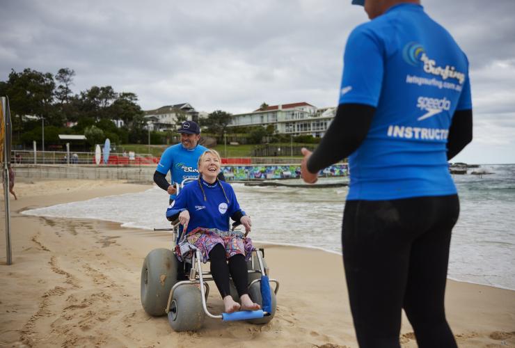 新南威尔士州，悉尼，邦迪海滩，一位坐轮椅的女士在沙地上接受“海滩冲浪之旅”教练指导 © 澳大利亚旅游局版权所有