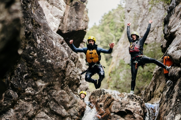 塔斯马尼亚州，摇篮山，在以摇篮山峡谷为背景的小瀑布顶端，有两个身穿救生衣、头戴安全帽的人，一人从岩石上跳下，另一人在欢呼 © 澳大利亚旅游局版权所有