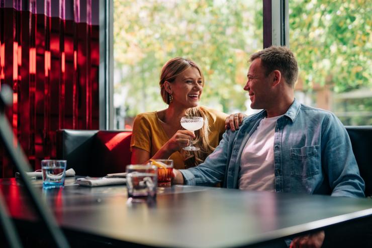 塔斯马尼亚州，霍巴特，一对夫妇在 Frank 餐厅手拿鸡尾酒微笑对望，身后是红墙 © Osborne Images 版权所有