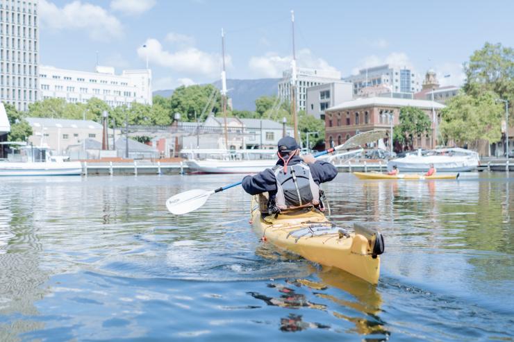塔斯马尼亚州，霍巴特，参加 Roaring 40s Kayaking 之旅的一位游客在霍巴特海滨平静的水域中划皮艇，前方有帆船和建筑物 © Wing Yin Ng 版权所有