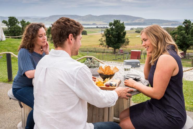 塔斯马尼亚州，科尔河河谷，煤河农场，三人围坐在桌旁享用熟食拼盘，背景是围场风光和河流 © Alastair Bett 版权所有