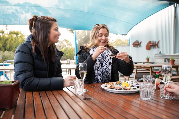 塔斯马尼亚州，大湾区，Get Shucked，蓝色顶棚下，两名女性坐在桌前，吃着一大盘生蚝搭配气泡酒 © Daniel Tran 版权所有