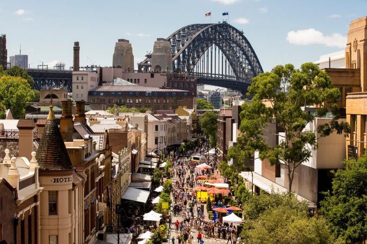 新南威尔士州，明媚阳光下，人们漫步在花伞林立的街道上的鸟瞰图，背景是悉尼海港大桥 © 新南威尔士州旅游局版权所有