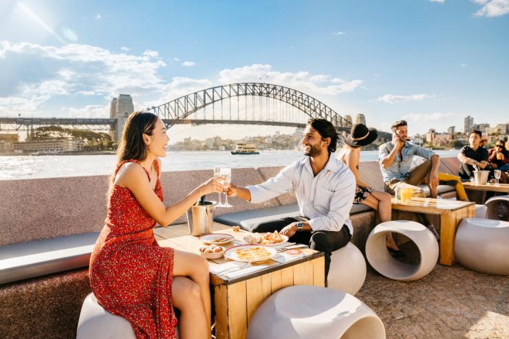 新南威尔士州，一位男士和一位女士在悉尼歌剧院酒吧享用气泡酒和美食并欣赏悉尼海港大桥的景色 © 新南威尔士州旅游局版权所有