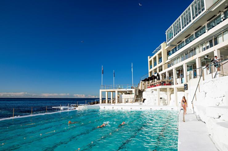 新南威尔士州，邦迪，人们在邦迪冰山泳池的碧水中游泳，左侧是海洋，右侧是亮白色的邦迪冰山俱乐部建筑 © 新南威尔士州旅游局版权所有