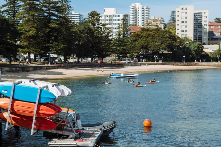新南威尔士州，曼利镇，一排橙色的皮划艇沿着曼利镇皮划艇中心岸边一字排开，周围人们在划皮艇 © 新南威尔士州旅游局版权所有