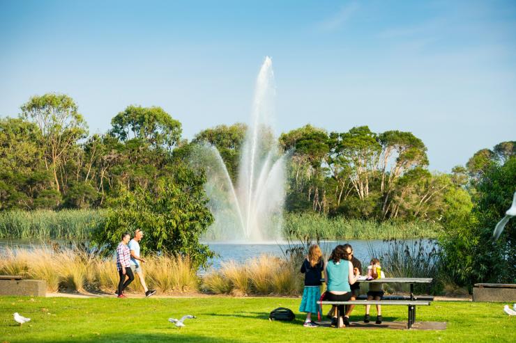 维多利亚州，赛尔，一家人正坐在草地上的野餐桌旁，同时有两人沿着 Lake Guthridge 漫步，湖中央有一座水景装置 © Rob Blackburn 版权所有