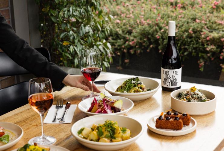 维多利亚州，摩林顿半岛，Jackalope Rare Hare Wine &amp; Food Store，一只人手伸向餐桌上的一杯红葡萄酒，桌上摆满了现做美食 © Amy Whitfield 版权所有