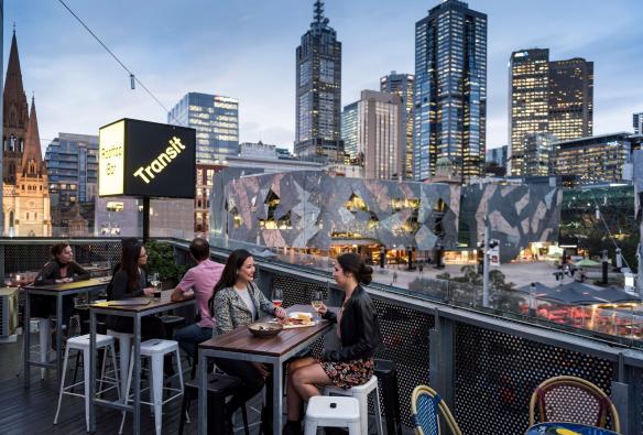 维多利亚州，墨尔本，两位女性在 Transit Rooftop Bar 享用饮料，背景是墨尔本的市区景观 © 维多利亚州旅游局版权所有