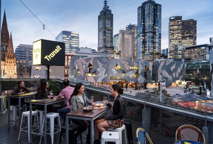 维多利亚州，墨尔本，两位女性在 Transit Rooftop Bar 享用饮料，背景是墨尔本的市区景观 © 维多利亚州旅游局版权所有