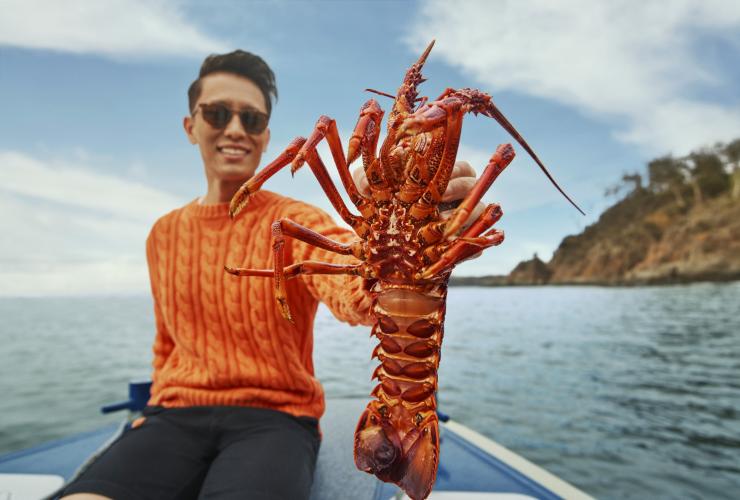 塔斯马尼亚州，布鲁尼岛，潘尼戈自然之旅，海鲜饕餮之旅，一个人坐在船上，手持龙虾对着镜头 © 澳大利亚旅游局版权所有