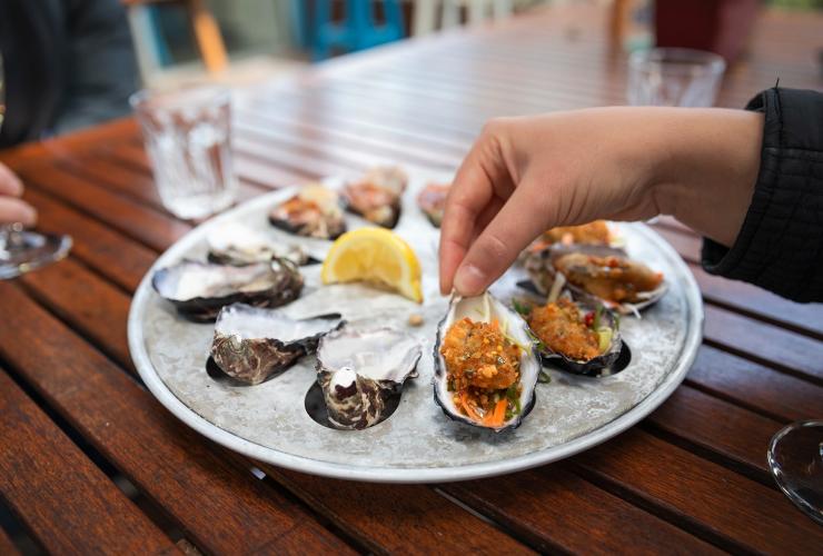 塔斯马尼亚州，布鲁尼岛，Get Shucked 餐厅，一盘铺在冰上的生蚝和将一只培根焗生蚝举起的一只手的特写 © 澳大利亚旅游局版权所有