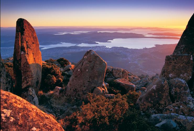 塔斯马尼亚州，惠灵顿山/kunanyi 山顶，前景中是被朝阳映红的岩石，远处是霍巴特和海洋的景色 © 塔斯马尼亚旅游局/Garry Moore 版权所有